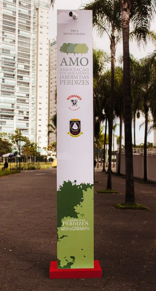 Totem da Associação de Moradores com câmera de monitoramento, na calçada arborizada do bairro