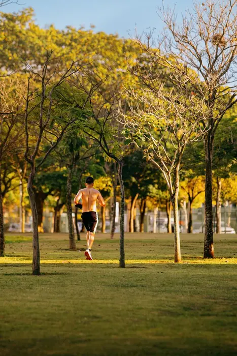 Morador correndo no gramado do parque, entre árvores, com o sol baixo ao fundo