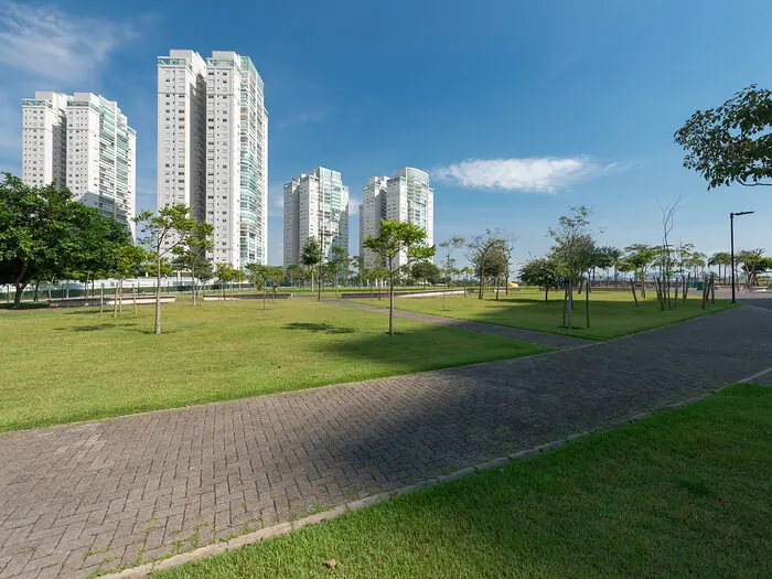 Alameda de piso intertravado atravessando o gramado do parque, com as torres ao fundo e um poste de iluminação sem nenhum fio