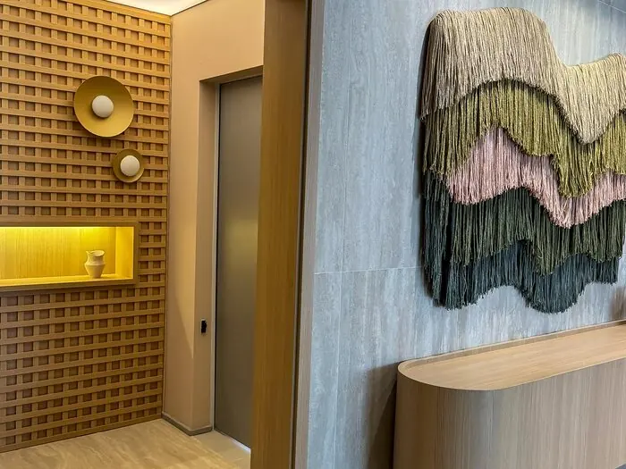 Hall privativo com porta de elevador entre uma parede de madeira gradeada com nicho iluminado e uma parede de pedra com painel de fibras e aparador curvo